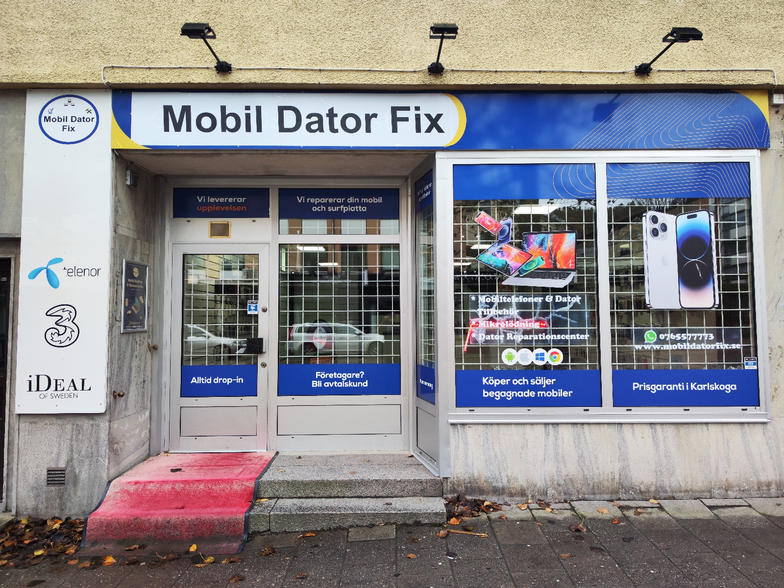Fasad till "Mobil Dator Fix" med skyltar som erbjuder reparationer, försäljning och service av mobiler och datorer på svenska. Entrén har en rosa ramp, gallerförsedda fönster och olika företagslogotyper på den vänstra väggen. Kontakta oss för mer information.